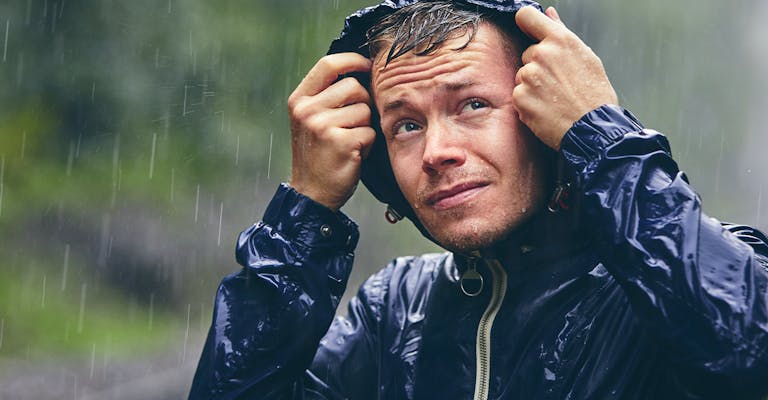 Portrait d'un jeune homme en tenue de pluie avec une capuche, trempée sous une pluie battante. Son regard est orienté vers le ciel.